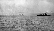 Four warships at sea sailing in line ahead, the shoreline distant in the background