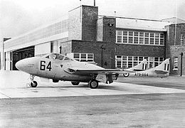 Side view of a single-engined, twin-boomed military jet parked in front of a building