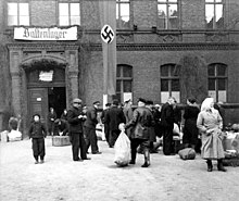 Schwarzweißfotografie eines Gebäudes mit der Aufschrift „Baltenlager“ und der Hakenkreuzflagge. Im Vordergrund sammeln sich Menschen mit Koffern und Säcken.