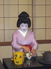 A geisha wearing a pink kimono sat at a black table, whisking a small cup of tea.
