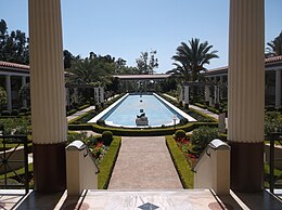 A long and rectangular fountain, with a gray statue in it, at the center of a courtyard. The fountain is flanked by shrubbery, trees, and flowers; around the perimeter of the courtyard is a building with white pillars and mahogany roofs.
