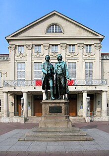 Photograph of a large bronze statue of two men standing side by side and facing forward. The statue is on a stone pedestal, which has a plaque that reads "Dem Dichterpaar/Goethe und Schiller/das Vaterland". Behind the monument there is a large, 3-storey building with an elaborate stone façade.