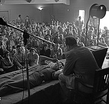 A mostly seated crowd watches as Hubbard, seated on a chair, speaks to a woman lying prone in front of him.