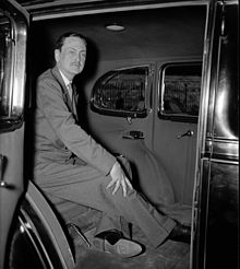 Man in sack suit sits in a car, hand on his splayed right leg.