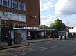 A red-bricked building with a blue sign reading "COLINDALE STATION" in white letters and several people in front all under a blue sky with white clouds