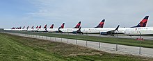 Many in-line airplanes with the Delta Air Lines logo on the tail, parked on the pavement behind a fence.