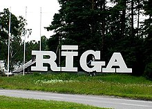 Frontale Farbfotografie von großen, weißen Buchstaben, die den Namen „Riga“ erzeugen und auf einer Wiese stehen. Auf dem i ist ein Strich, wie in der lettischen Orthografie. Im Hintergrund stehen Bäume.