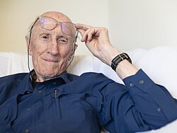close-up of Stewart Brand wearing a dark blue shirt, holding his glasses up slightly above his eyes, smiling and looking left of camera