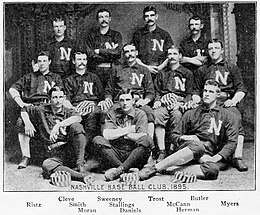 A black and white photograph of twelve men arranged in three rows, standing, sitting in chairs, and sitting on the floor. They are wearing dark baseball uniforms with a white "N" on the chests.