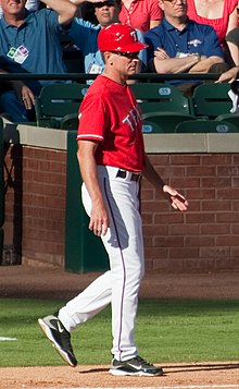A man wearing a red batting helmet and baseball jersey with white pants walking on a baseball field