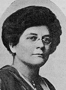 Middle-aged white woman wearing glasses, her dark hair arranged in an updo.
