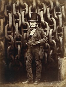 A 19th-century man wearing a jacket, trousers, and waistcoat, with his hands in his pockets and a cigar in mouth, wearing a tall stovepipe top hat, standing in front of giant iron chains on a drum.