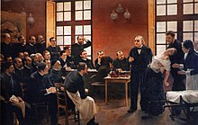 A painting of a 19th-century medical lecture: At the front of the class, a woman faints into the arms of a man standing behind her, as another woman, apparently a nurse, reaches to help. An older man, the professor, stands beside her and gestures as if making a point. Two dozen male students watch them.