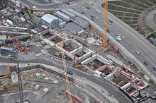 A construction site with several cranes and walls surrounding a long, rectangular hole in the ground.