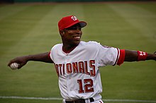 With baseball in hand, an African-American man wearing a white and red Nationals baseball uniform cocks his arm backward as he prepares to throw.
