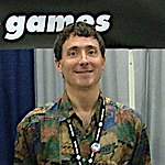 A Caucasian man with short brown hair and a convention pass on a lanyard around his neck smiles for a camera. Part of the Telltale Games logo is visible in the background.