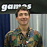 A man wearing a multicolored shirt smiles at the camera. Behind him stands a blue and white background, above which is an excerpt of logo which reads "games".