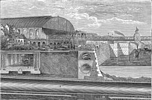 Illustrated section showing below ground structures of a riverside embankment including a covered railway line with steam train, riverside wall with sewer and pipes running behind. A large, glass roofed railway station sits in the left middle distance adjoining a railway bridge that crosses the river. Boats ply the water and tiny figures are engaged in construction work with a raised scaffold in the centre.