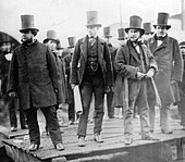 A group of ten men in nineteenth-century dark suits, wearing top hats, observing something behind the camera