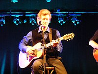 Singer Rodney Crowell, seated on a stool and strumming an acoustic guitar.