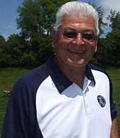 A smiling white tanned man with grey hair, wearing sunglasses and a blue-collared white shirt, outside on a playing field.