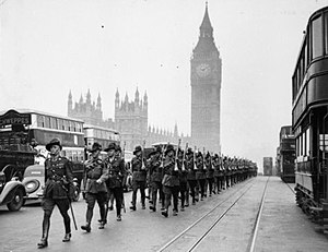 Black and white photograph of a large number of men wearing military uniforms marching down a street. A tower with a clock on it is visible in the background.