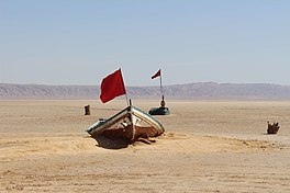 Photograph showing a flat sandy landscape. In the foreground a red flag flies from a broken old boat. Behind it, another red flag is anchored in the ground by a solid looking piece of metal machinery. The flags may be position markers for times with more water.