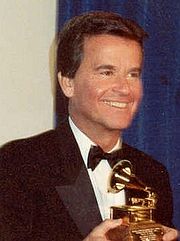 Headshot of a television host in a black suit and bowtie holding a small trophy depicting a gilded gramophone.