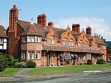 A row of brick cottages with a turret at each end and four shaped gables between them. One of the central houses is decorated with hanging baskets and other floral arrangements.