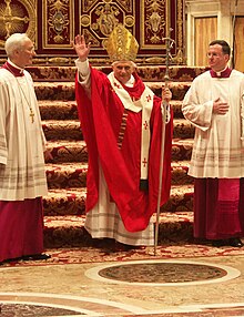 Frontale Farbfotografie vom Papst in rotweißer liturgischer Kleidung mit goldener Kopfbedeckung. Er trägt einen Kreuzstab und hält seinen rechten Arm zum Segen. Neben ihm steht jeweils ein Würdenträger. Im Hintergrund sind von Teppich bedeckte Stufen zu sehen.