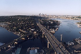 A freeway with a smaller bridge next to it spanning a waterway to a wooded but still urbanized hillside with a city's skyline in the distance