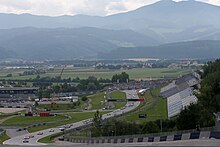 A tarmac course overlooked by a grandstand makes its way uphill through alpine meadows, with a large mountain in the background.