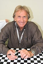 Derek Bell holding a biro pen in both of his hands is smiling at the camera