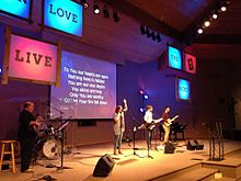 A contemporary worship team leads the congregation using lyrics projected on a motion background, and coordinated lighting.