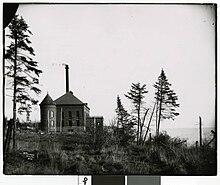 Black-and-white photo of a Romanesque revival building along a lakeshore with pine trees