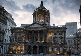 A neoclassical building in two storeys with a central portico and a dome seen almost end-on. It is flanked on each side by other buildings