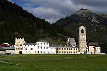 Frontale Farbfotografie einer Klosteranlage mit Bergen im Hintergrund. Mehrere Häuser stehen auf einer Alm nebeneinander und rechts ist eine Turmuhr.