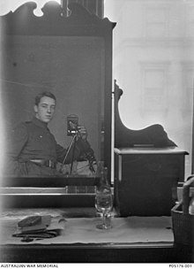 A dresser with a large mirror and a bottle of wine, a glass and some papers sitting on top. A man in military uniform can be seen in the mirror's reflection. He has his hand in his pocket, and is using a camera on a tripod.