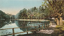 A colorized image of a path running along the bank of a lake and up towards a dirt road. The lake is still and glassy, and trees and a fence rest along its banks.