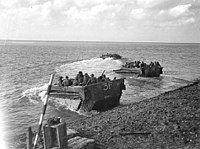An amphibious vehicles carrying troops drives up out of the water onto a beach. A similar vehicle is yet to reach the shore