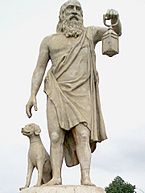 Stone statue of a bearded man in ancient Greek dress holding a lantern. A sculpted dog sits at his side.