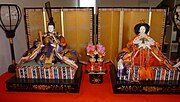 An Emperor doll with an Empress doll, in front of a gold screen. The optional lampstands are also partially visible.