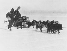 Two men in polar gear drive a sledge dog team pulling a heavily laden sledge