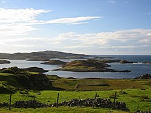 A view from a low eminence looking towards a fence and a tumble-down stone wall beyond which is a strip of uncultivated land and the sea shore. A variety of hummocky green and brown islands hug the coast as waves line their rocky shores under a blue sky.
