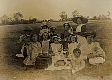 monochrome photograph of early 20th century group of thirteen adults and children seated with a picnic on a grass slope with hedge and trees in the background