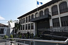 This is a picture showing the building of the Constitutional Court of Guatemala. It shows a two story building colonial Spanish type, with multitude of windows and a reddish tile roof, with a background of a blue sky on top. In the foreground there is a white sculpture depicting Pegasus, next to a concrete pathway leading to the entrance of the building. There are three police standing on the sidewalk apart from each other, in guard duty. There is also a couple of large banners between two of the police in the right of the picture, about the mission of the Court and recommending people to respect the integrity of the building.