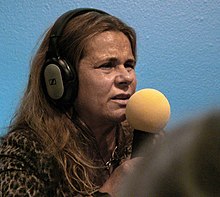 Porträt einer Frau mit Kopfhörern, die in ein gelbes Mikrofon spricht. Die Frau hat halblange dunkelblonde Haare, die sie offen trägt. Sie steht vor einer blauen Wand.