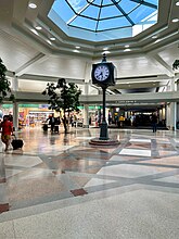 An open rotunda space with indoor trees, a large skylight above, and a freestanding clock in the center. Airline passengers walk by with luggage.