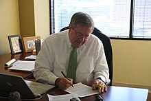 A grey-haired man with a white shirt and green tie sits at a desk in front of an open window, looking down to follow his pen as he writes on a sheet of paper