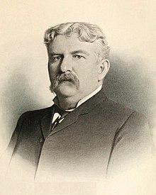 Black and white photograph of a man with a long mustache and short wavy hair wearing a black suit